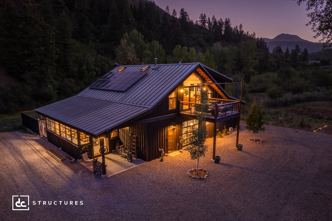 A cozy, illuminated two-story cabin with a metal roof sits in a wooded landscape at dusk. Solar panels are visible on the roof. Surrounding trees and a mountain are silhouetted against a purple evening sky.