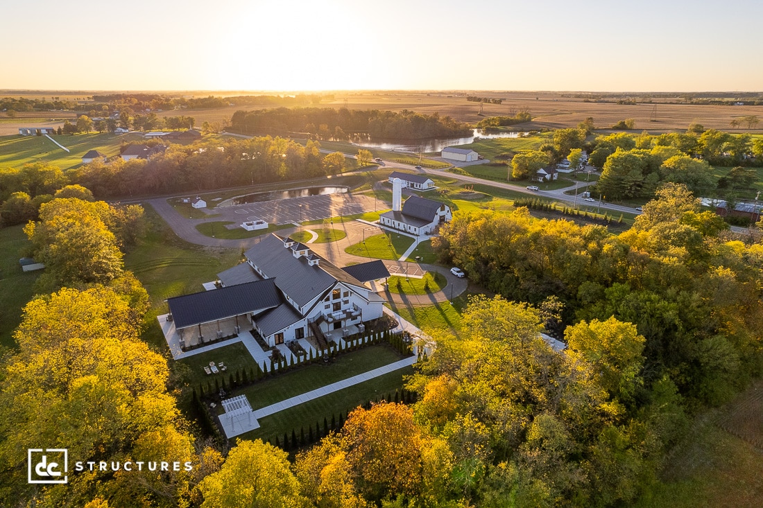 Aerial view of a picturesque rural landscape at sunset, featuring several white buildings with black roofs amidst lush green trees. Surrounding fields stretch into the distance, under a clear blue sky tinged with golden hues.