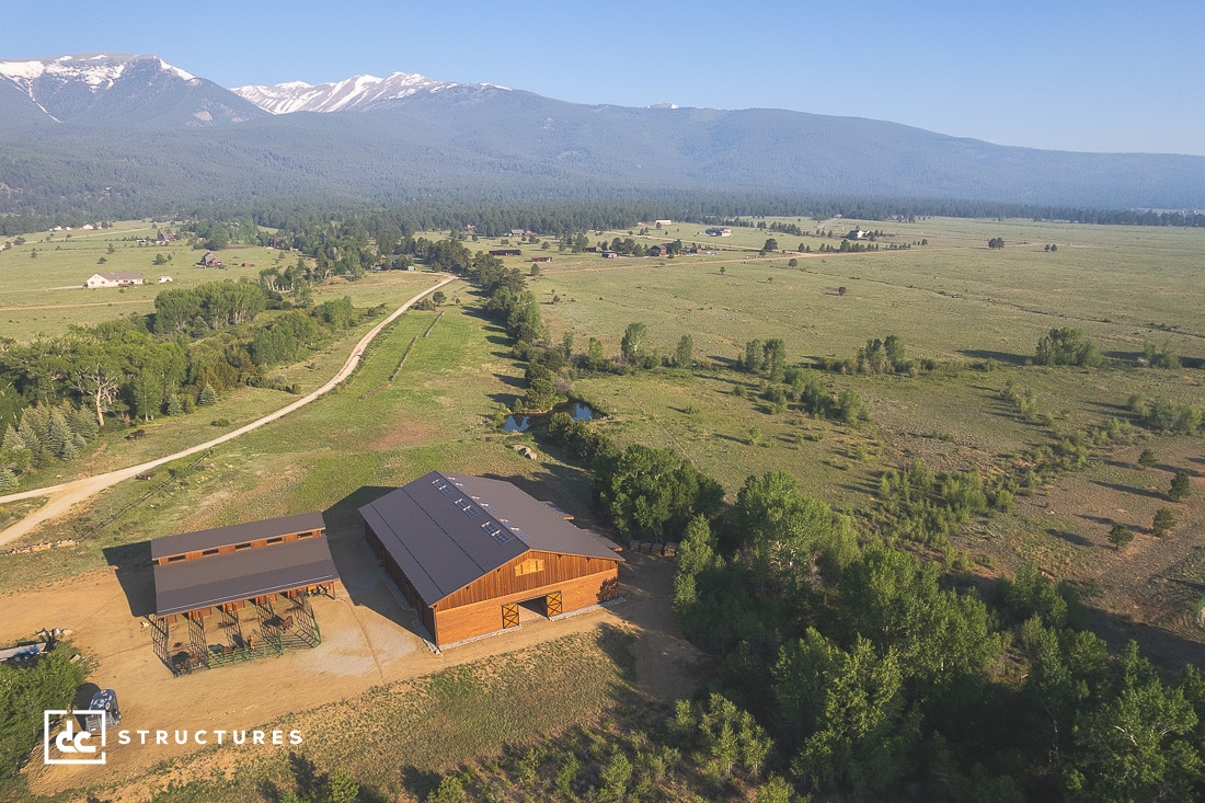 Aerial view of a large wooden barn and adjacent structure set in a vast grassy landscape. A dirt road leads to the buildings, surrounded by trees. Mountains with patches of snow are visible in the distance under a clear blue sky.
