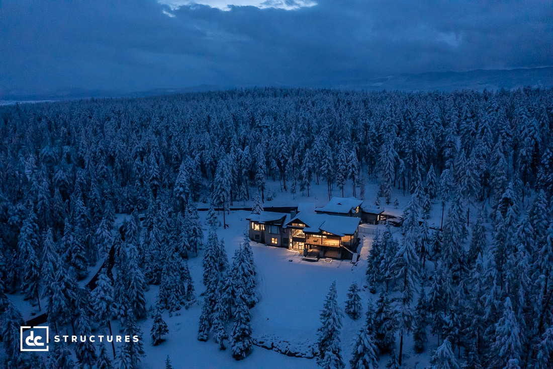 A cabin with lit windows stands surrounded by a dense forest blanketed in snow. It's twilight, casting a blue hue over the snowy landscape, creating a serene winter scene.