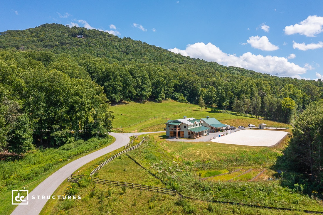 A scenic, aerial view of a large property with a green-roofed building and a gravel area surrounded by lush green hills and trees under a bright blue sky with fluffy clouds. A winding road leads to the property.