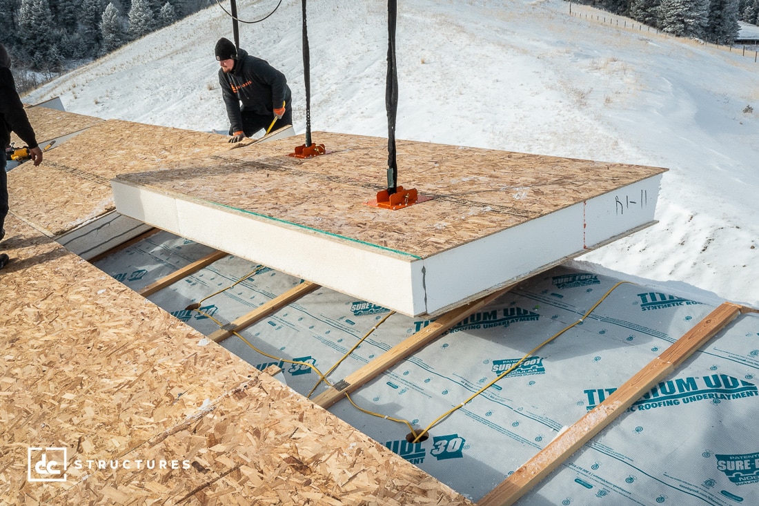 Workers installing a large prefabricated insulated panel on a roof with a snowy landscape in the background. The panel is being lifted by a crane, and the construction area is covered with protective material.