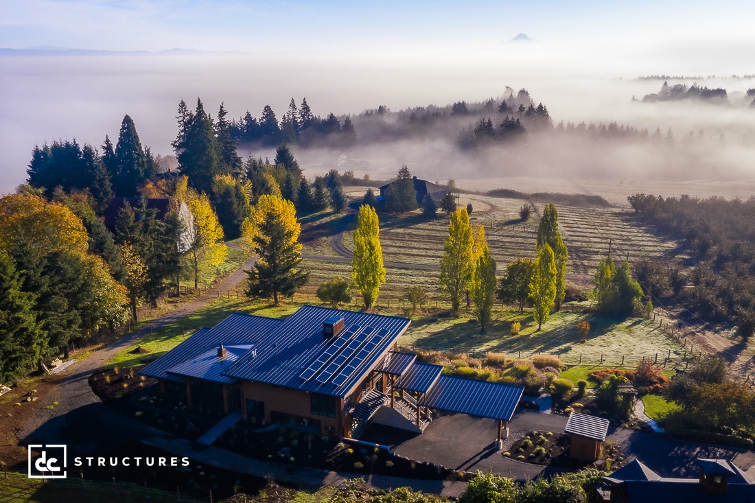 Aerial view of a modern house with a solar panel roof surrounded by trees and fields, with morning fog and distant mountains in the background.