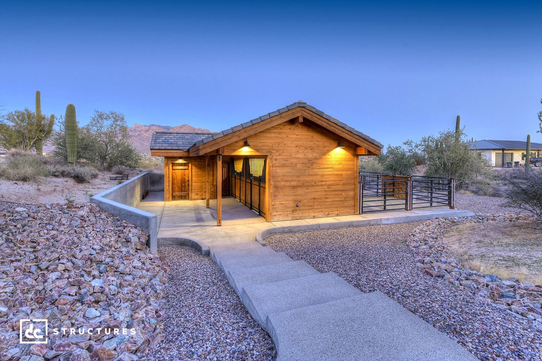 Single-story wooden house with a sloping roof, warmly lit entrance, and surrounding desert landscape. Features a pathway leading to the front door, with cacti and mountains in the background.