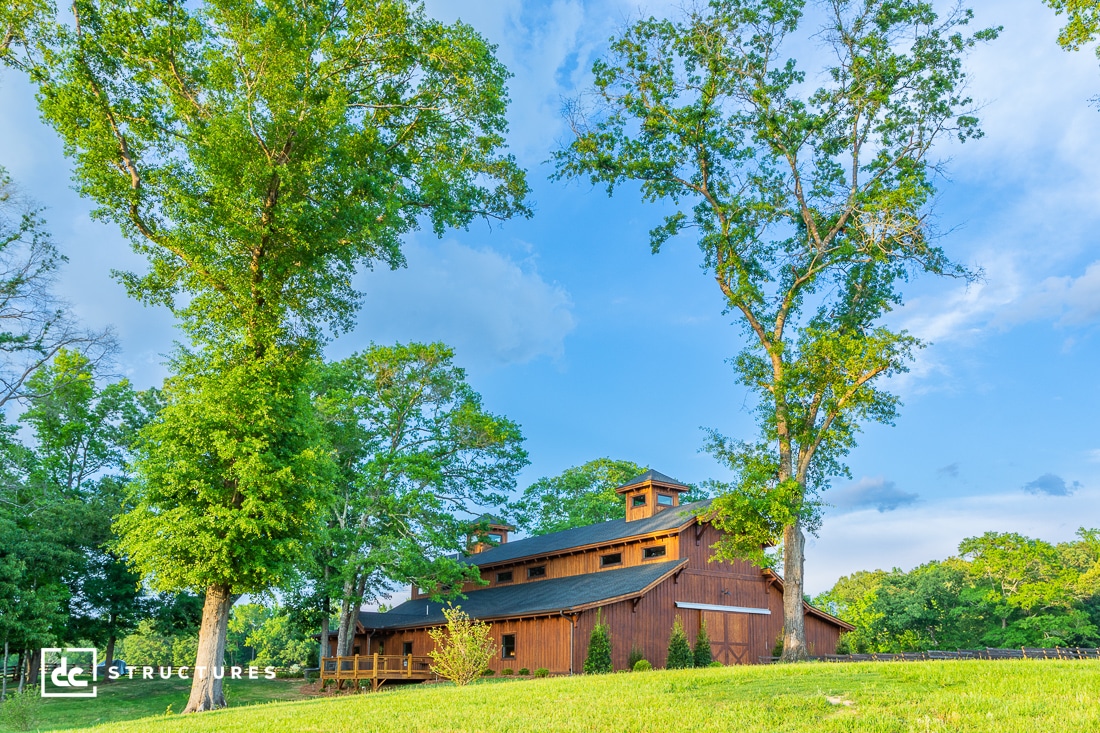 A rustic wooden barn is surrounded by tall, leafy trees under a blue sky with scattered clouds. The grass is lush and green, creating a picturesque rural scene.