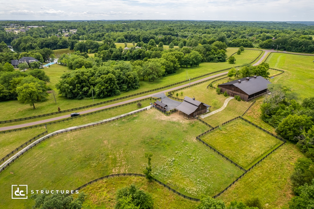 Aerial view of a rural property with a large barn and farmhouse surrounded by green fields and trees. A winding road leads to the buildings, and fenced areas are visible. The landscape extends to the horizon under a partly cloudy sky.