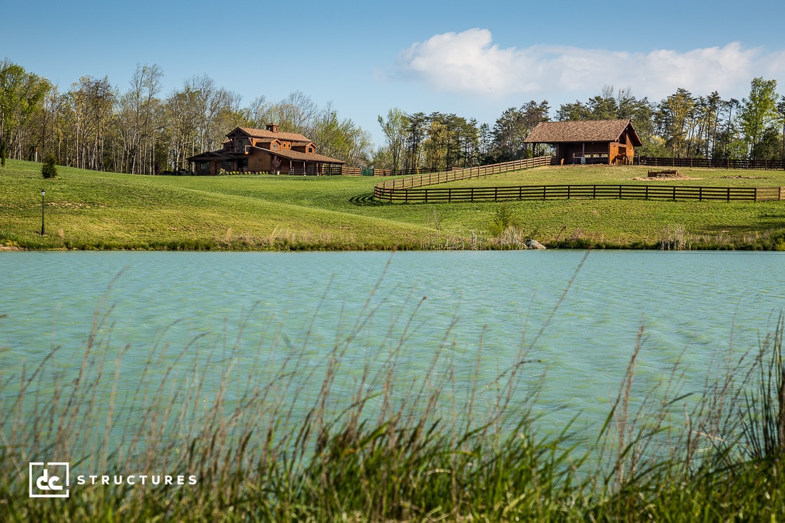 Rustic wooden barns on a lush green hillside overlooking a tranquil pond, surrounded by trees under a clear blue sky. A wooden fence runs through the landscape, emphasizing the pastoral setting.