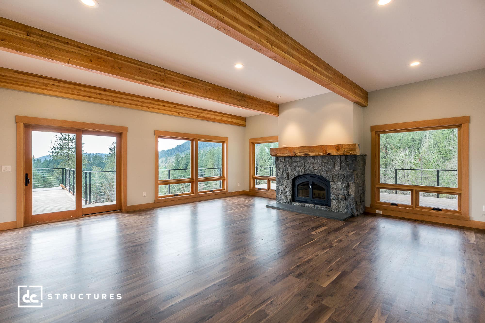 Spacious living room with wooden beams, large windows, and double doors leading to a balcony. A stone fireplace is centered on the back wall. Hardwood floors complement the natural, forested view outside.