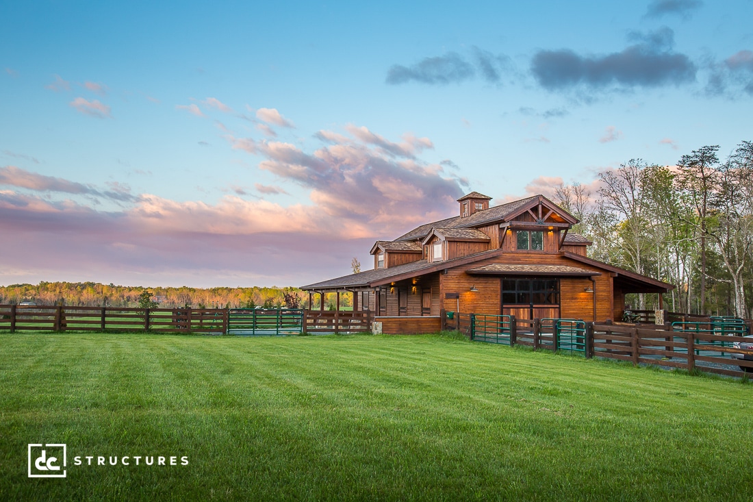 A wooden barn-style house sits on a lush green field, surrounded by a wooden fence. The sky is blue with scattered clouds, and the sun casts a warm glow on the building. Trees are visible in the background.