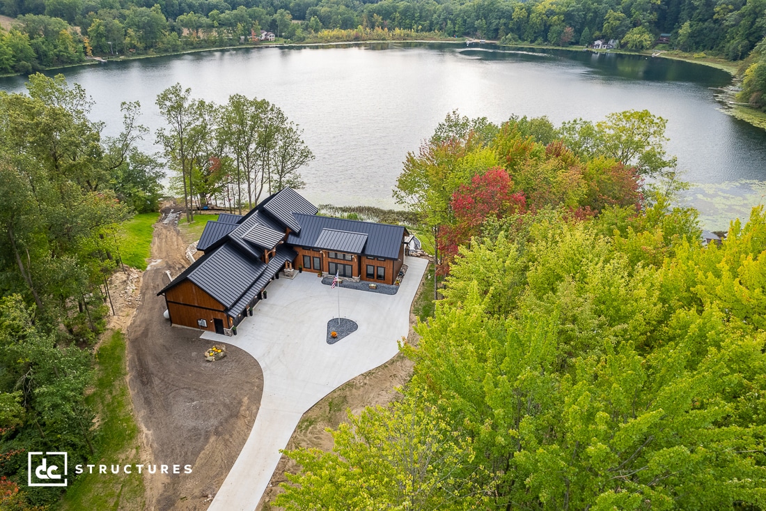 Aerial view of a modern house with a black metal roof, surrounded by lush green trees and a few buildings. The property is near a large, tranquil lake. A winding driveway leads to the house, and a small patio area is visible.