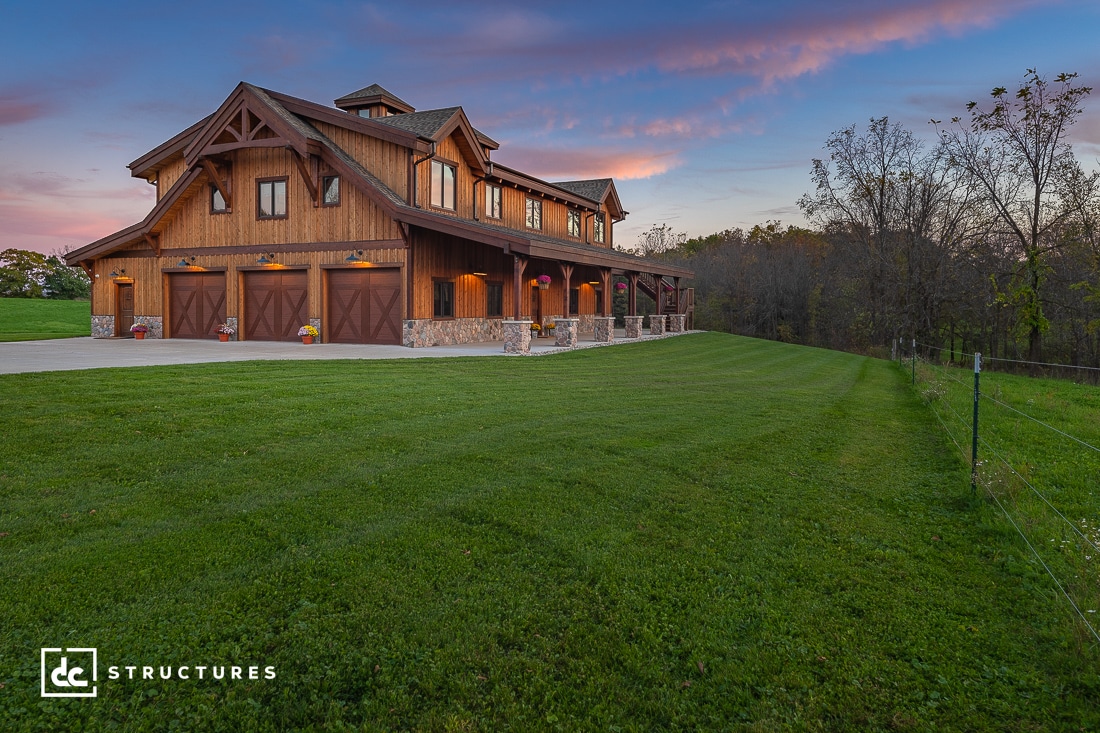 A rustic two-story wooden house with stone accents, surrounded by lush green grass. The sky is a vibrant mix of pink and blue at sunset. A line of potted flowers decorates the porch. Trees are visible in the background.