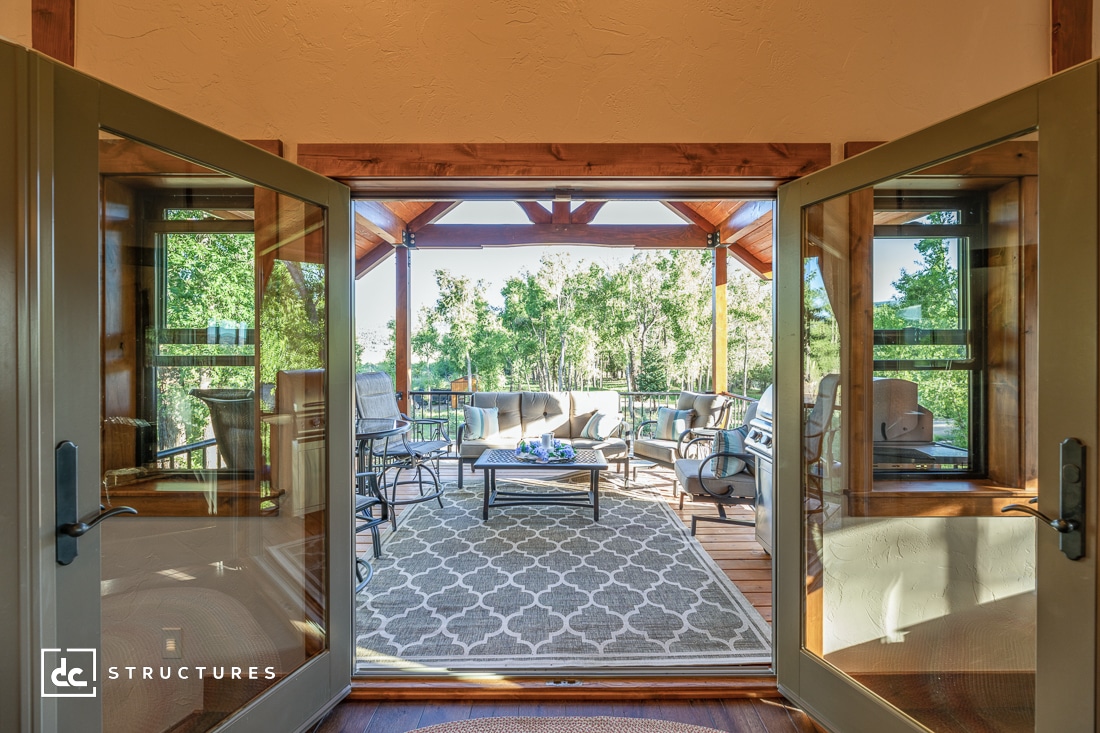 Open double doors leading to a sunlit outdoor patio with seating, a patterned rug, and surrounded by trees. Wooden beams frame the patio area.