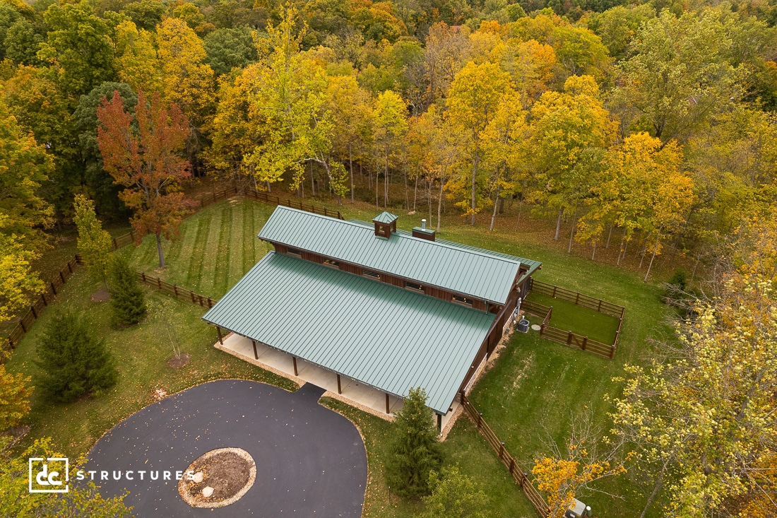 Aerial view of a large metal-roofed barn surrounded by trees with autumn foliage. The barn sits behind a circular driveway on a grassy area, enclosed by a wooden fence. The scene is set in a forested area with a mix of green and yellow leaves.