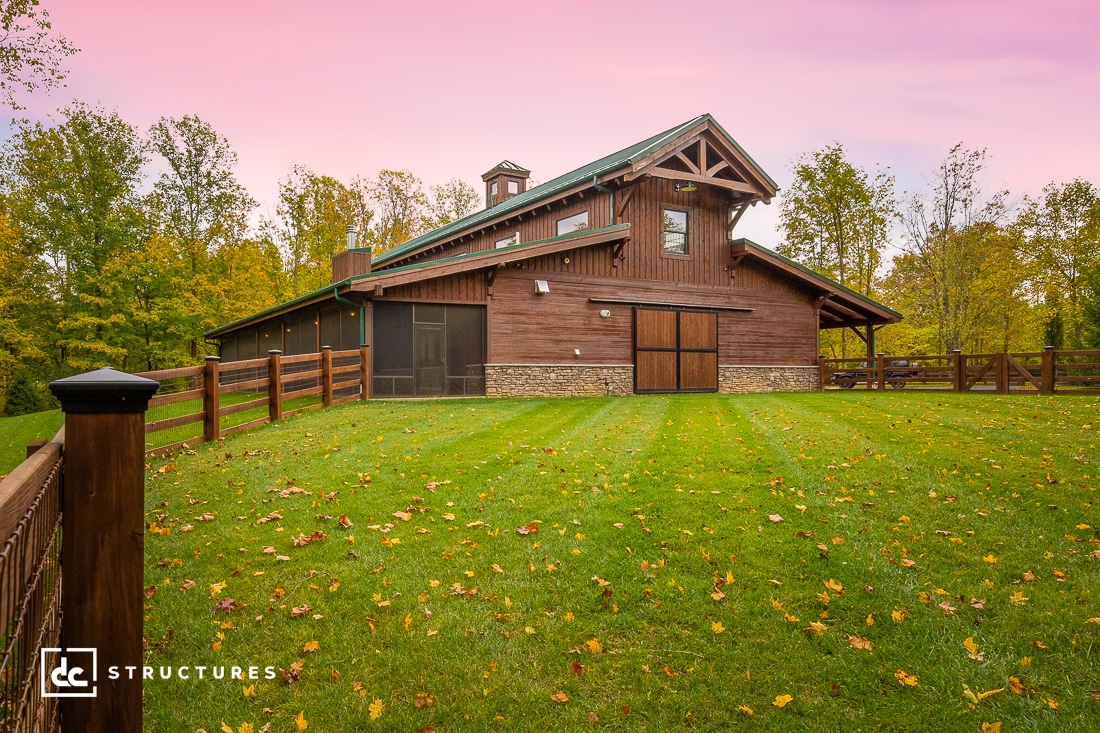 Wooden barn with a steep gabled roof, surrounded by a wooden fence and lush green lawn. Autumn leaves are scattered on the grass. Trees with early fall foliage surround the structure, and the sky has a pinkish hue.