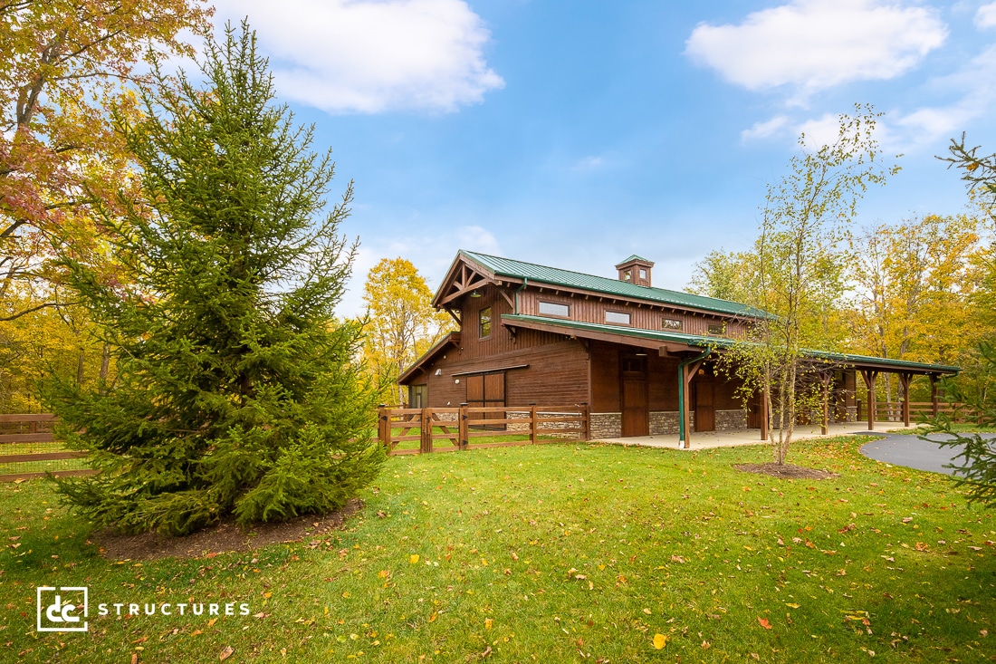 A picturesque wooden house with a green roof surrounded by lush green grass and trees. The sky is blue with scattered white clouds. A paved path leads to the entrance.