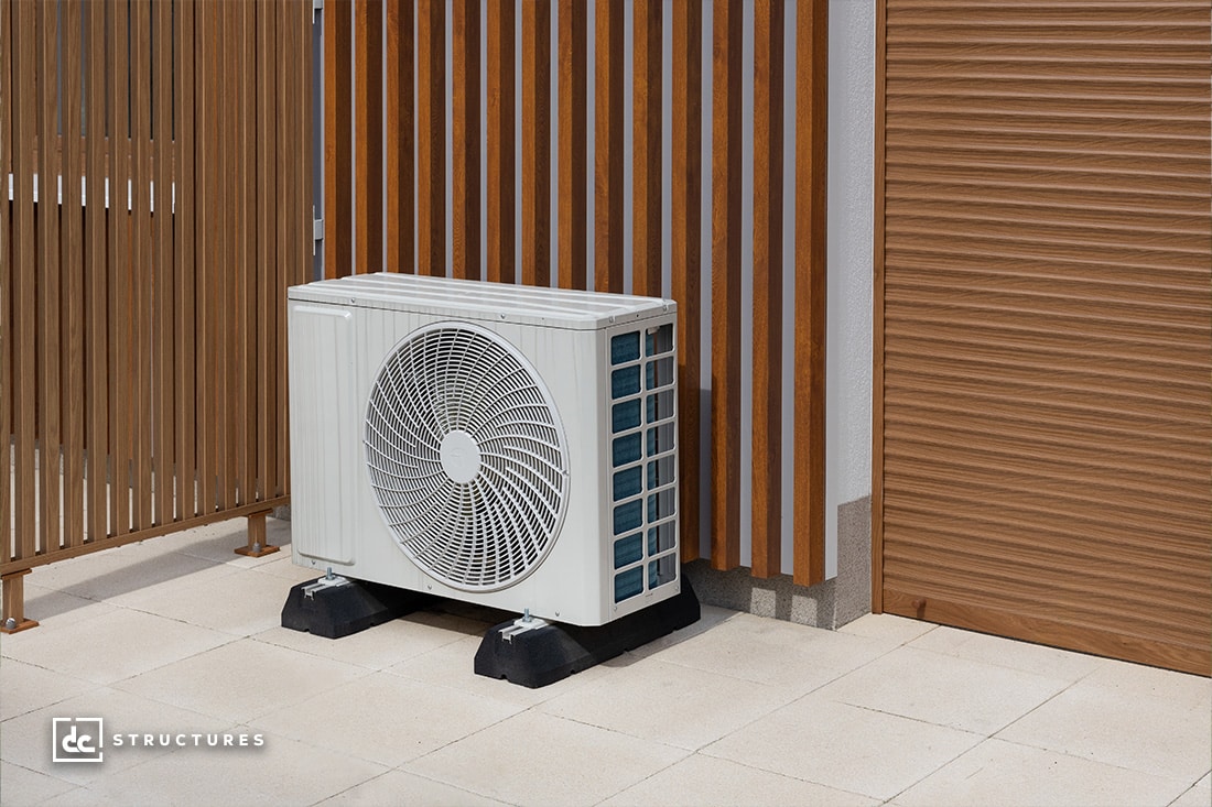 Outdoor air conditioning unit on a patio with wooden slat fencing and a closed wooden shutter in the background. The area is paved with light-colored tiles.
