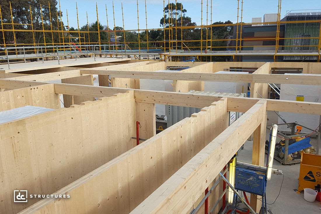 Construction site featuring a partially built wooden framework for a building, with exposed beams and scaffolding visible. Construction materials and equipment are scattered around the site, and there's a background of trees and neighboring structures.