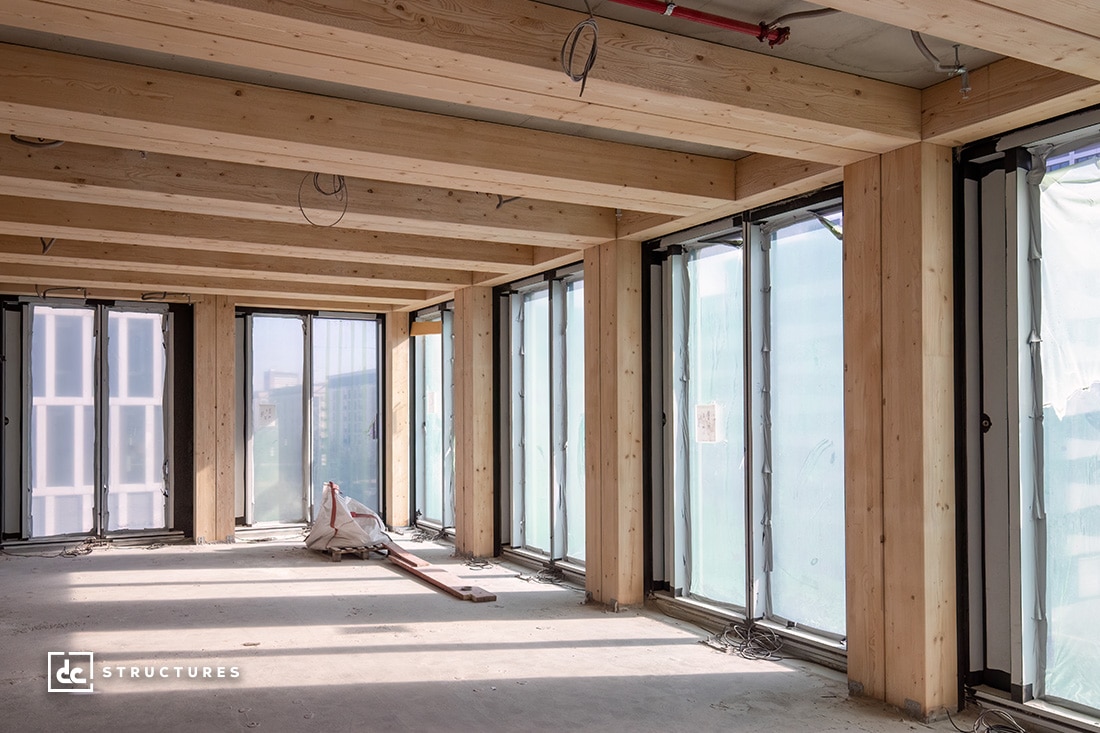 A sunlit, partially constructed building interior with large glass windows, wooden beams, and columns. Transparent plastic sheets cover the windows. Building materials and a metal tool are on the floor.