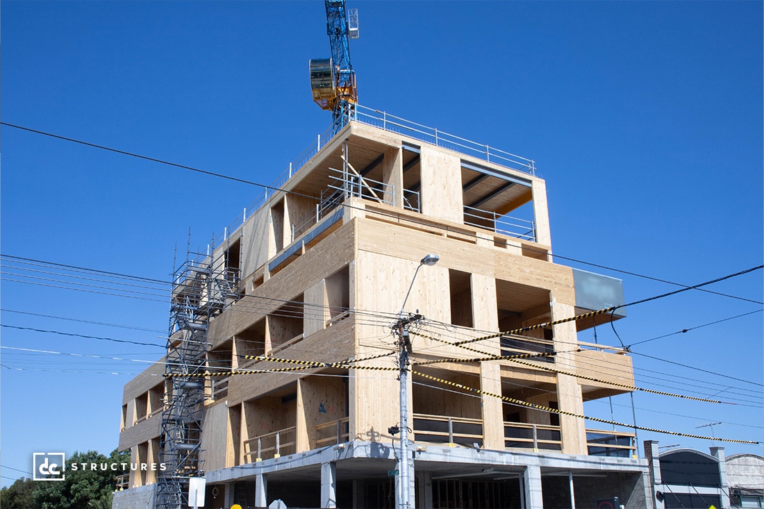 A multi-story wooden building under construction with scaffolding on the left side. A blue crane extends from the roof. The sky is clear and blue, and a few power lines cross in front of the structure.