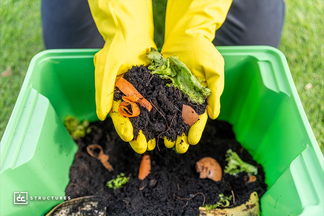 Person wearing yellow gloves holding compost with vegetable scraps and soil above a green bin. Grass is visible in the background.