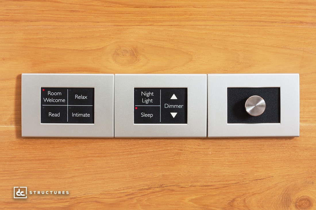 A wall-mounted control panel with three sections on a wooden backdrop. The left section has buttons for Room Welcome, Relax, Read, and Intimate. The middle has Night Light, Sleep, and Dimmer. The right section has a silver dial.