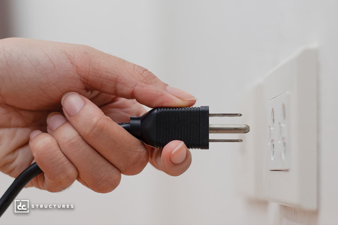 A person with medium-toned skin is plugging a black electrical cord into a white wall outlet. The background is a plain wall.