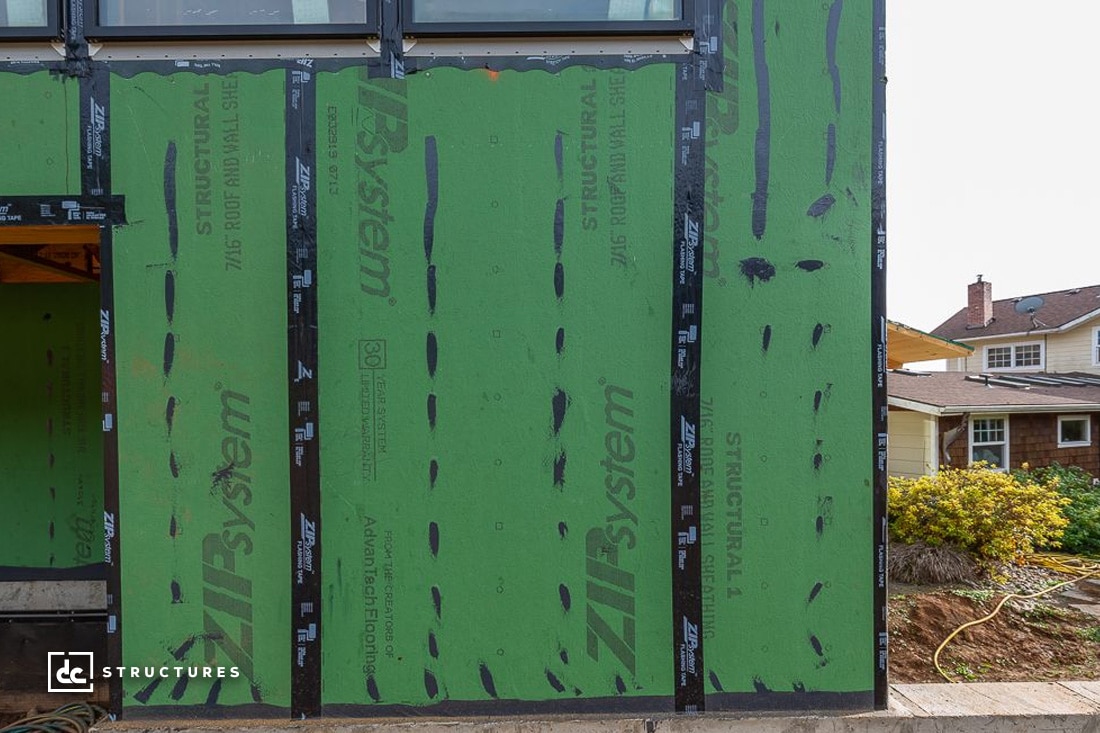 Partially constructed building with green sheathing panels on the exterior walls. A neighboring house is visible in the background, along with a garden area.