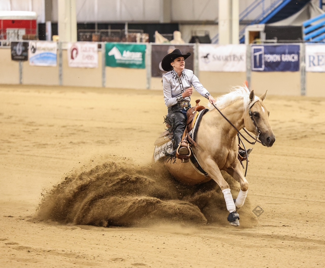 A cowboy in a black hat and striped shirt rides a light-colored horse, making a sharp turn on an indoor arena, kicking up a large cloud of sand as banners hang on the walls in the background.