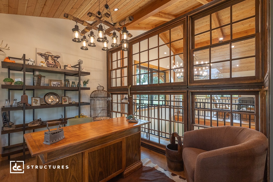 A rustic office with a wooden desk, brown armchair, open shelving with decor, and large interior windows overlooking a loft with exposed wood beams and railings. Warm lighting and natural wood give the space a cozy feel.