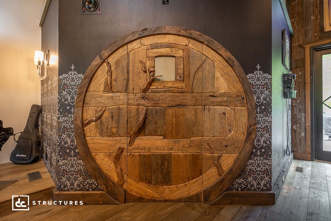 A large, round, wooden door with intricate carvings resembling a Hobbit-style entrance is set in a dark wall, flanked by patterned wallpaper and rustic wooden flooring.