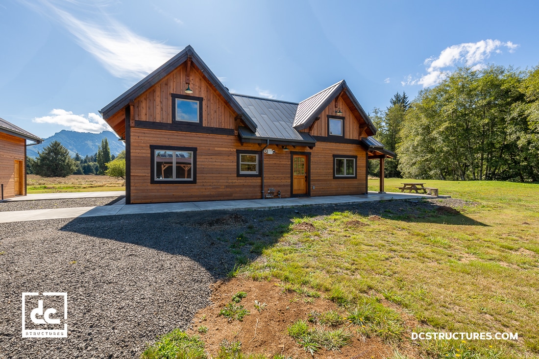 A two-story wooden cabin with a metal roof sits on a grassy lot, surrounded by trees and mountains under a clear blue sky. A picnic table is on the right, and the home has large windows and a side concrete path.