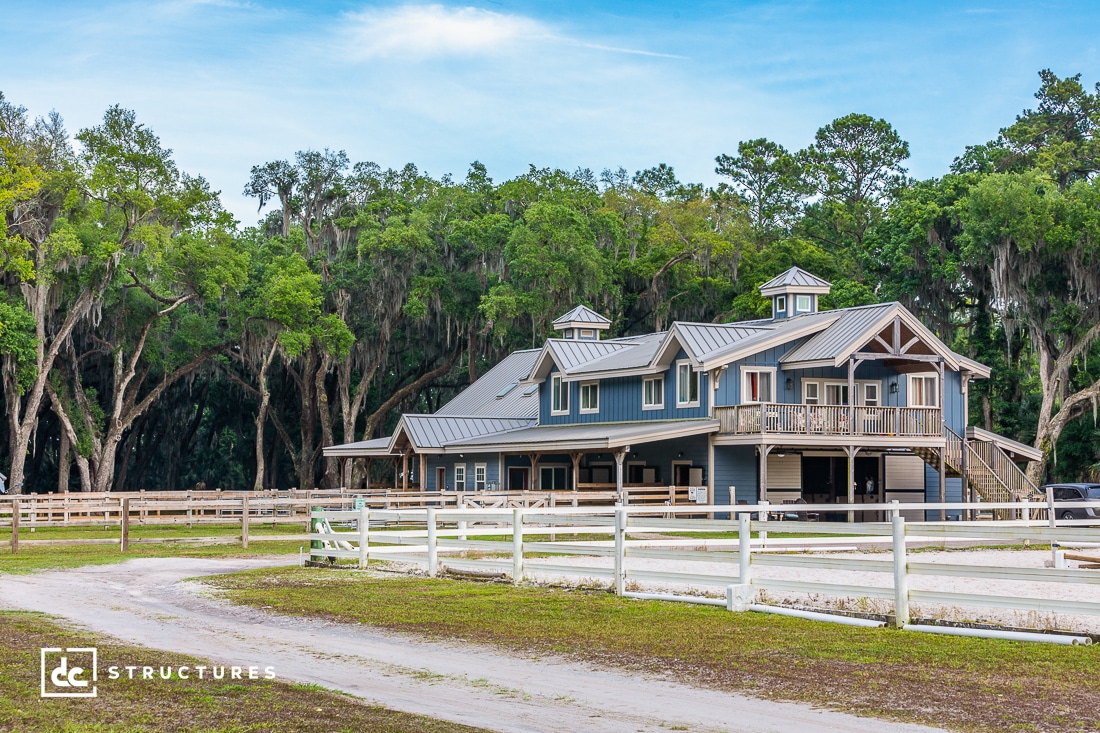 A large, modern barn with a metal roof and cupolas sits behind a white fence, surrounded by trees and greenery under a blue sky. A dirt road leads up to the structure, and the area appears well-maintained.