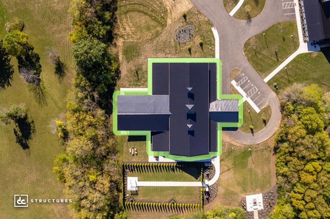 Aerial view of a large, dark-roofed building with green-highlighted edges, surrounded by trees, grass, parking areas, and walkways, photographed on a sunny day.