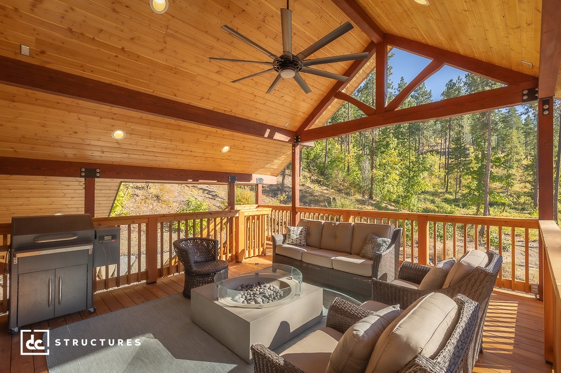 A covered wooden patio with wicker furniture, a fire pit table, and a grill. The ceiling has exposed beams, recessed lighting, and a large ceiling fan. Surrounded by trees and natural scenery.