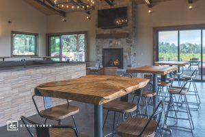 A modern rustic restaurant interior with wooden tables, metal chairs, a stone fireplace, large windows with scenic views, and a mounted TV above the mantel. The space is bright and inviting.