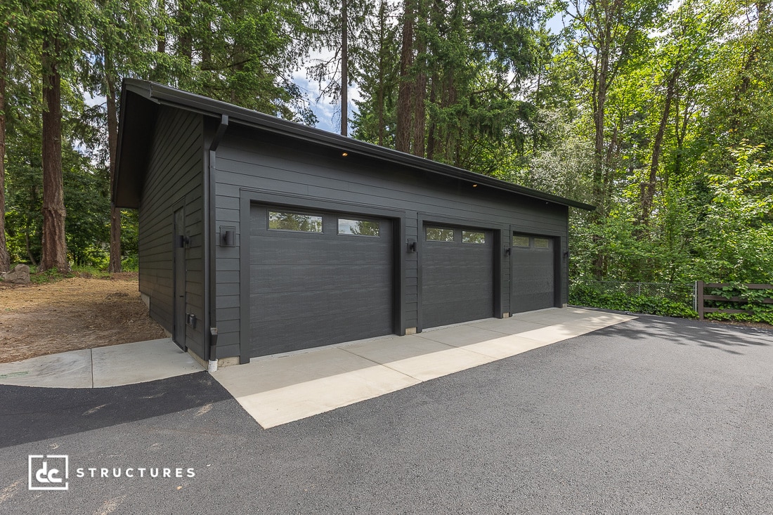 Modern, dark gray, three-car garage with flat roof and glass-panel doors, surrounded by tall trees and greenery, on a paved driveway.