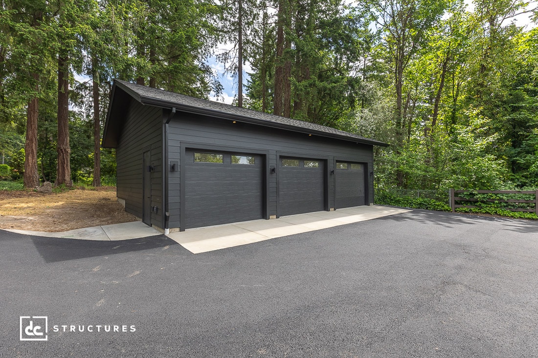 A modern, dark gray three-car garage with windows on each door sits beside a paved driveway, surrounded by dense, green forest and tall trees.