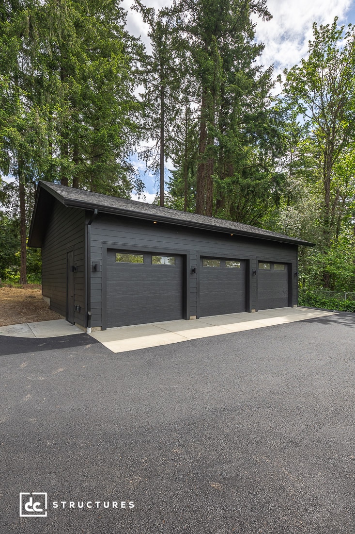 A modern, dark gray three-car garage with large windows on each door, surrounded by tall green trees and a newly paved driveway.