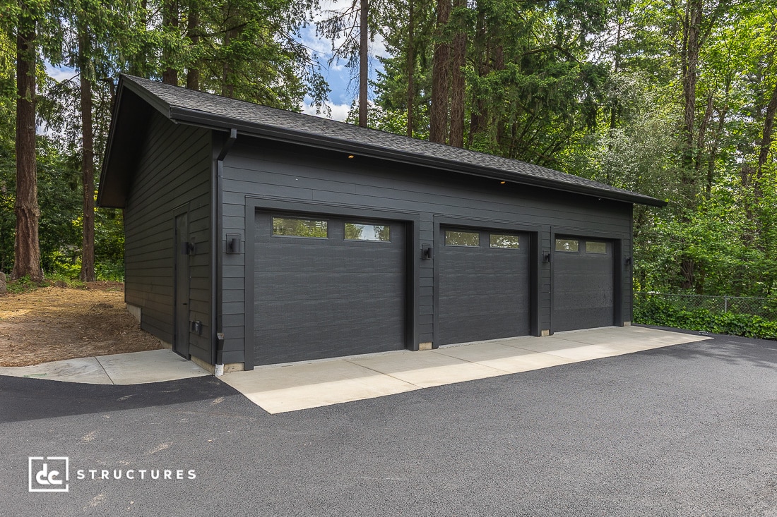 A modern dark gray three-car garage with glass windows on each door, surrounded by tall trees and set on a paved driveway. The structure has a clean, simple design and is nestled in a wooded area.