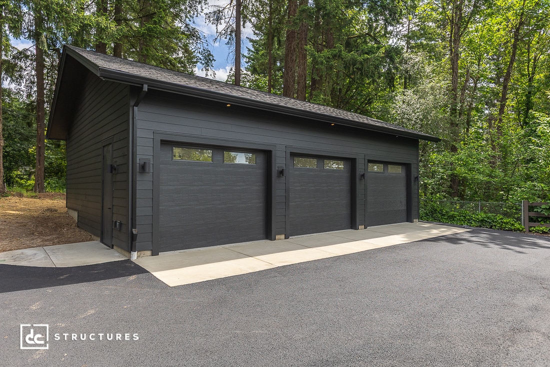 A modern three-car garage with dark gray siding and three separate doors, surrounded by trees and greenery, on a paved driveway.