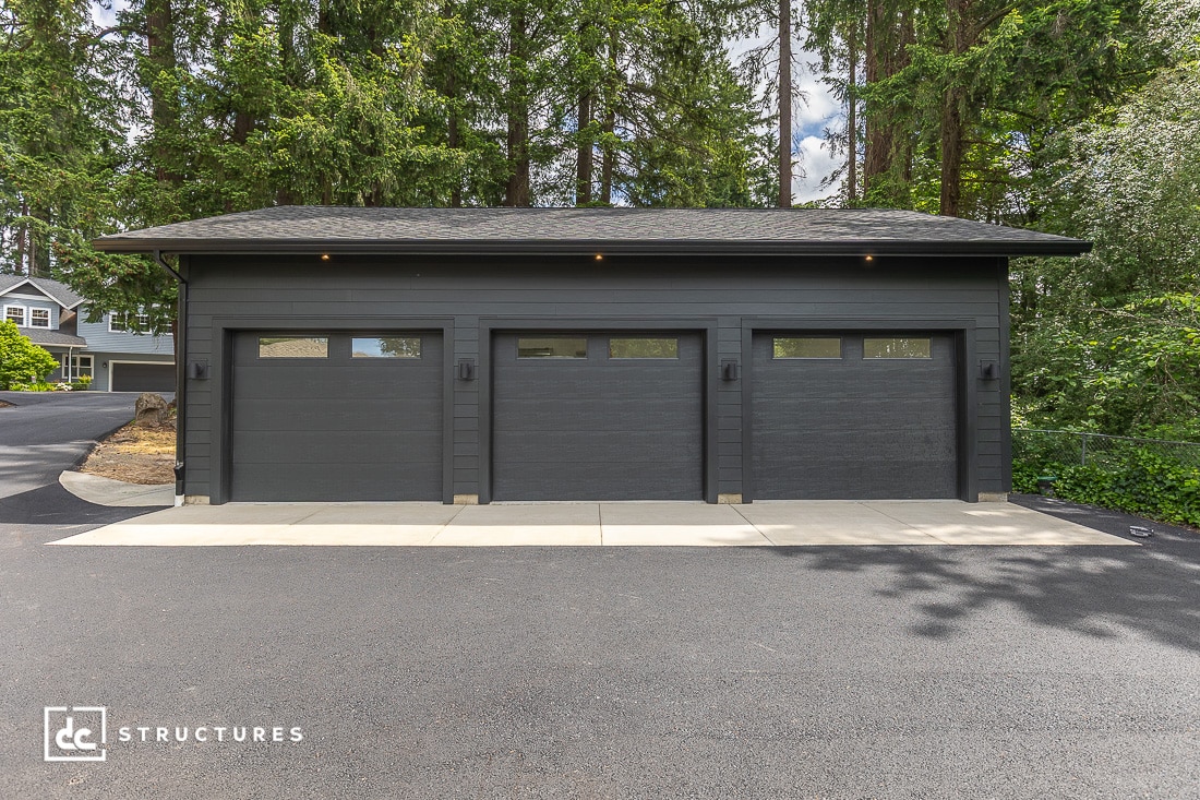 A modern dark gray three-car garage with three closed doors, set against a backdrop of tall evergreen trees and a paved driveway, with a house visible in the background.