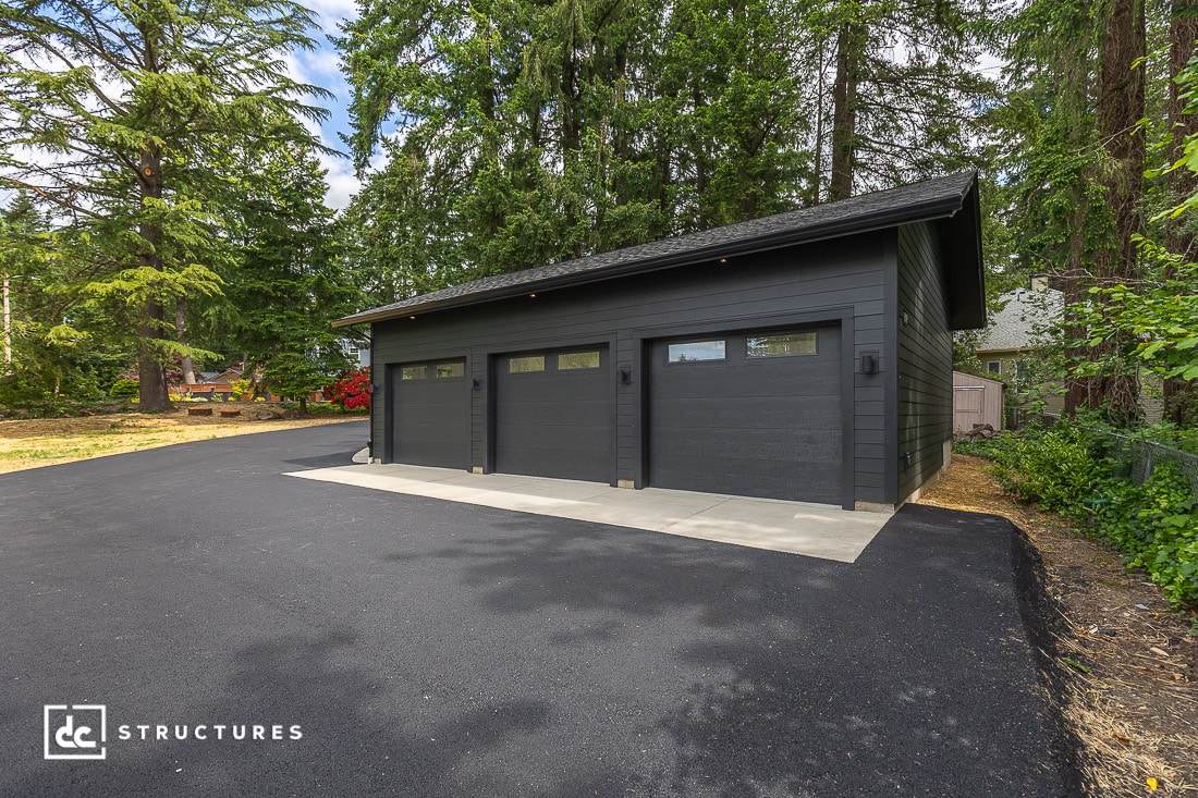 A modern, dark gray three-car garage with flat black doors sits on a freshly paved driveway, surrounded by tall green trees and vegetation.