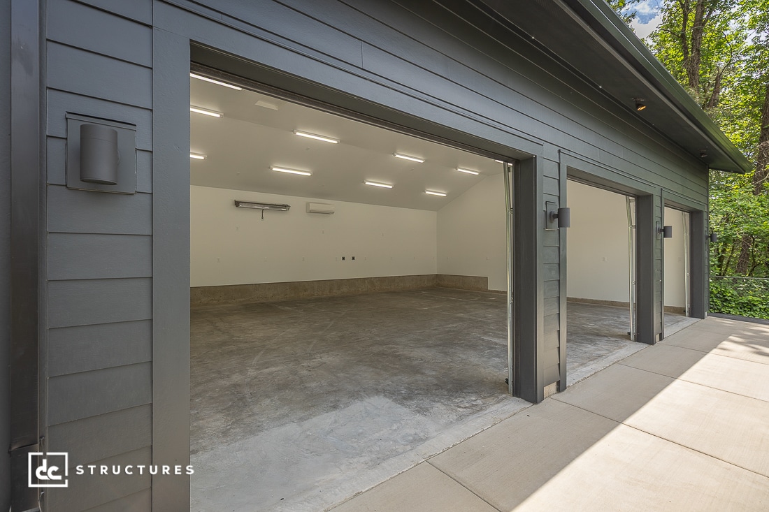 A modern three-car garage with open doors, gray exterior siding, bright interior lighting, and an empty concrete floor, surrounded by trees.