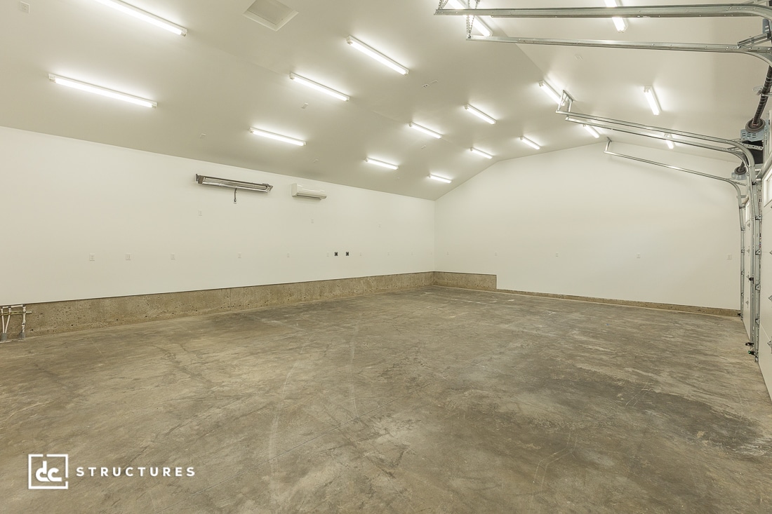 A spacious, empty garage with polished concrete floors, white walls, a high sloped ceiling, and bright overhead LED lights. An open garage door and HVAC units are visible on the back wall.