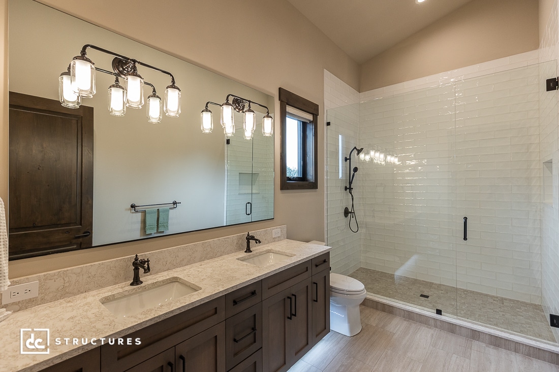A modern bathroom with a double-sink vanity, large mirror, wall-mounted light fixtures, a glass-enclosed shower, and a toilet. The decor features dark wood cabinets and neutral tile flooring.