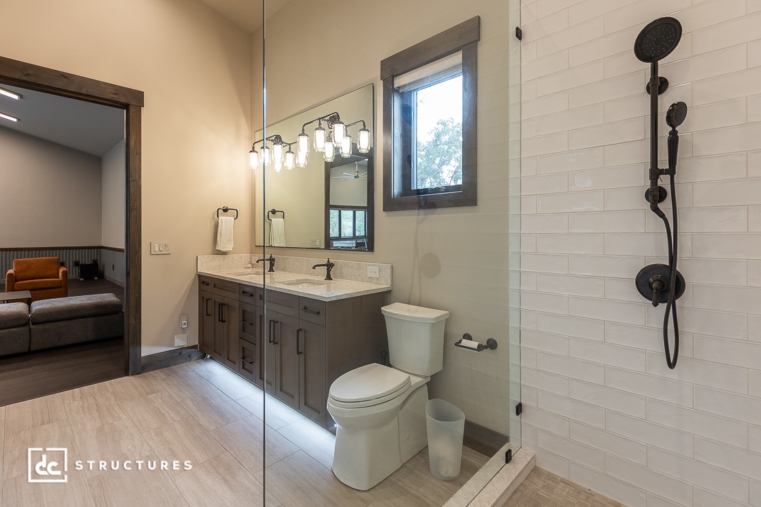 Modern bathroom with a double-sink vanity, large mirror, wall-mounted lights, toilet, glass-enclosed shower with black fixtures, window, and light wood floor. An adjacent room with a sofa is partially visible.