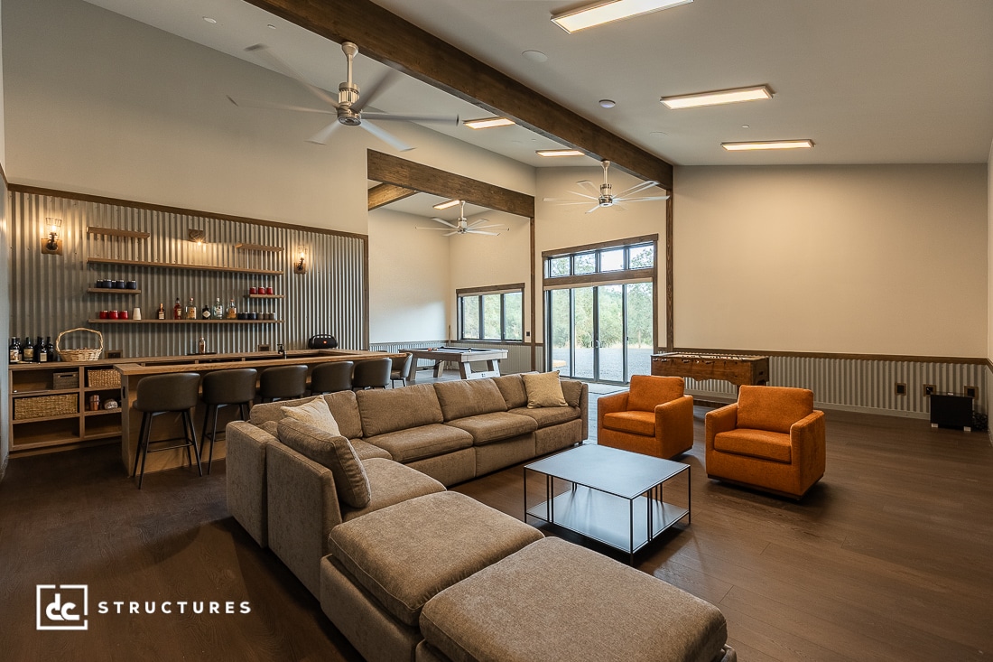 Modern lounge with a large sectional sofa, two orange chairs, a foosball table, and a bar area with stools. The room features wood floors, large windows, and ceiling fans.