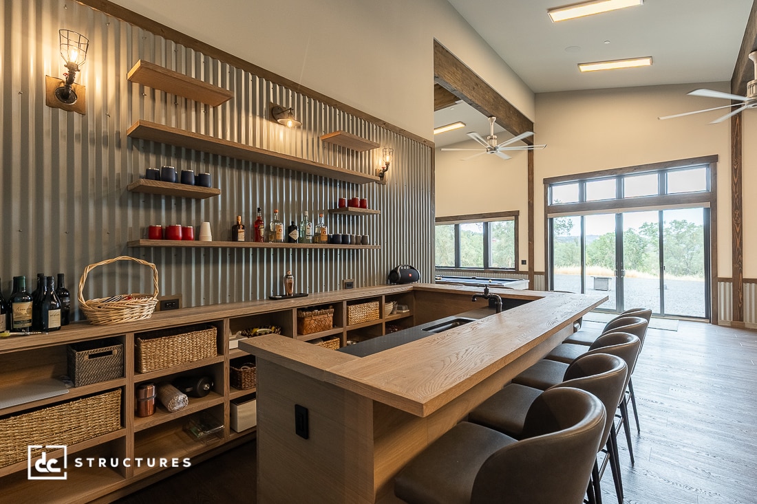 A modern home bar with a wooden counter, high-back chairs, open shelves holding bottles and cups, corrugated metal wall accents, and large windows letting in natural light.