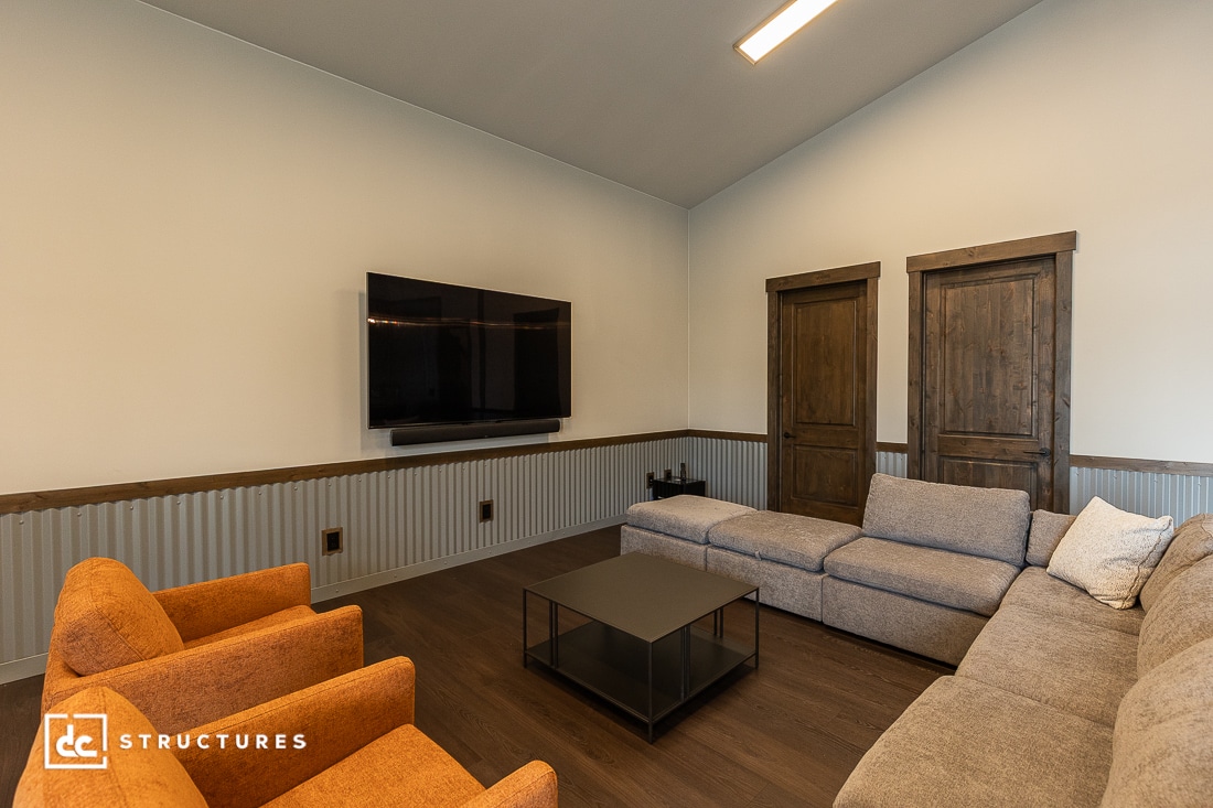 A modern living room with a gray sectional sofa, two orange armchairs, a black coffee table, a wall-mounted TV, wooden doors, and a corrugated wainscoting accent on the lower half of the walls.