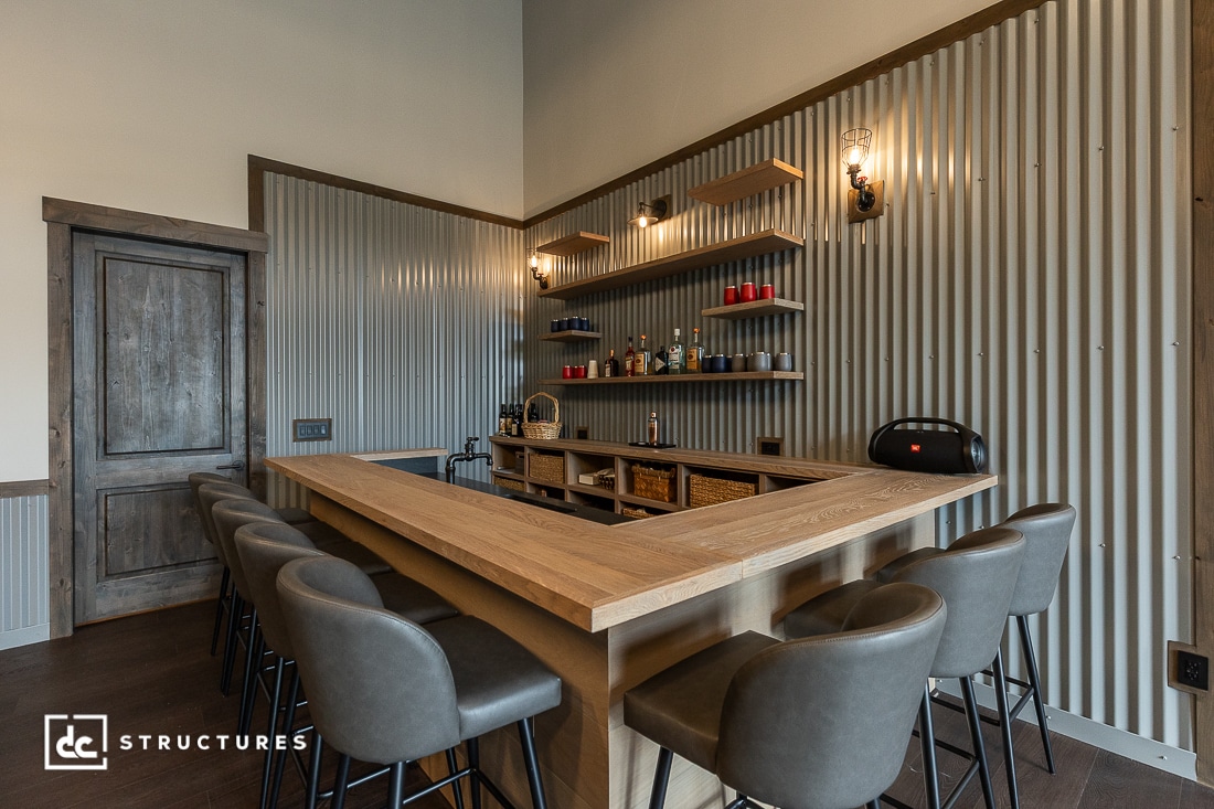 A modern home bar with a wooden U-shaped counter, six gray padded barstools, shelves with glasses and bottles, corrugated metal walls, and industrial-style lighting.